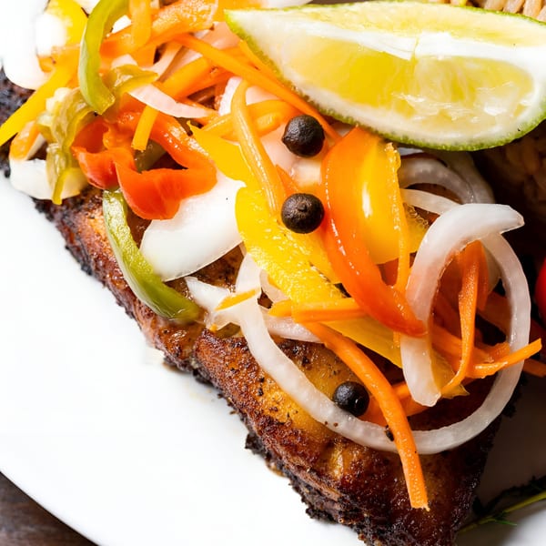 Close-up of Jamaican escovitch fish topped with colorful pickled vegetables and a lime wedge.