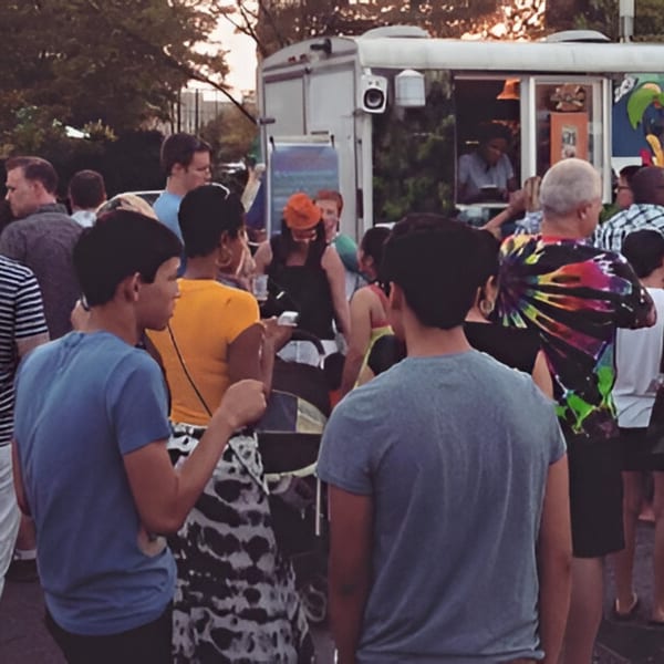 A crowd of people gathered around the Da Blue Lagoon Jamaican food truck at sunset.