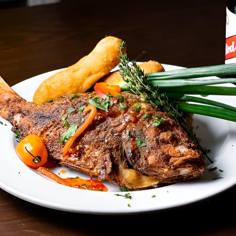 Whole fried fish with bammy, garnished with herbs, peppers, and scallions on a white plate.