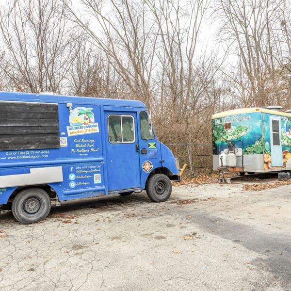 Two Da Blue Lagoon Jamaican Kitchen food trucks, one blue and one with a lagoon mural, parked outdoors.